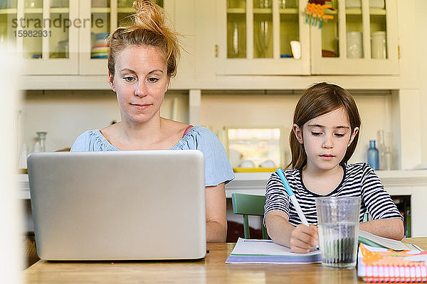 Frau arbeitet am Laptop und Mädchen (6-7) macht Hausaufgaben
