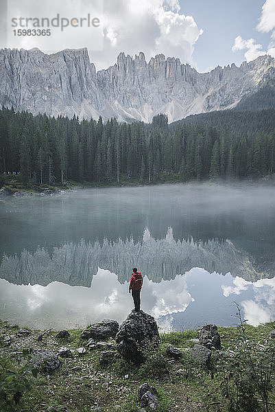 Italien  Carezza  Junger Mann steht in der Morgendämmerung auf Fels am Lago di Carezza in den Dolomiten