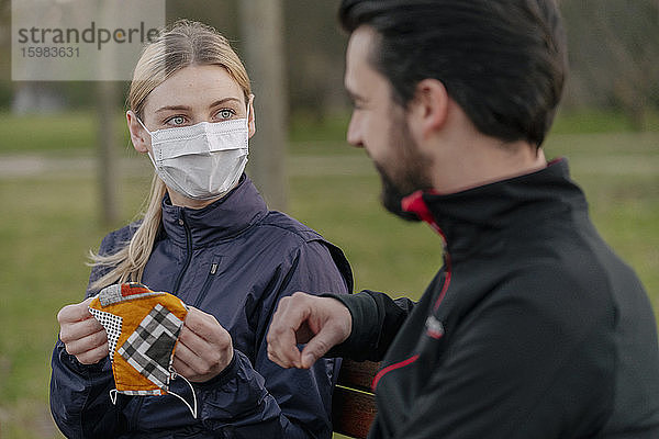 Junge Frau  die einem Mann während der Quarantäne im Park eine Gesichtsmaske gibt