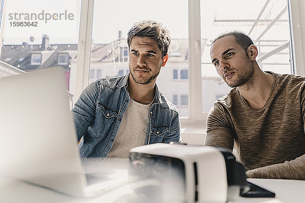 Zwei junge Geschäftsleute betrachten einen Laptop mit VR-Brille auf dem Schreibtisch