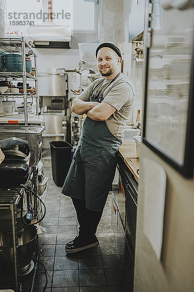 Lächelnder selbstbewusster männlicher Besitzer steht mit verschränkten Armen in der Küche eines Restaurants