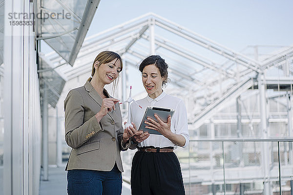 Zwei Geschäftsfrauen mit Tablet und Windradmodell bei einem Treffen in einem Gewächshaus