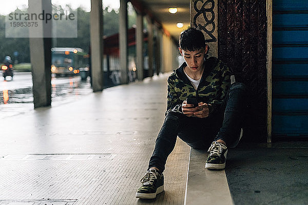Junger Mann benutzt sein Smartphone  während er auf einer Treppe an der Wand sitzt