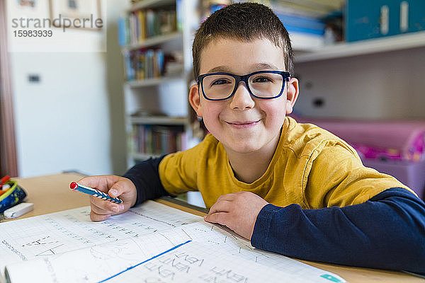 Lächelnder Junge sitzt mit einem Buch am Schreibtisch während des COVID-19 Hausunterrichts