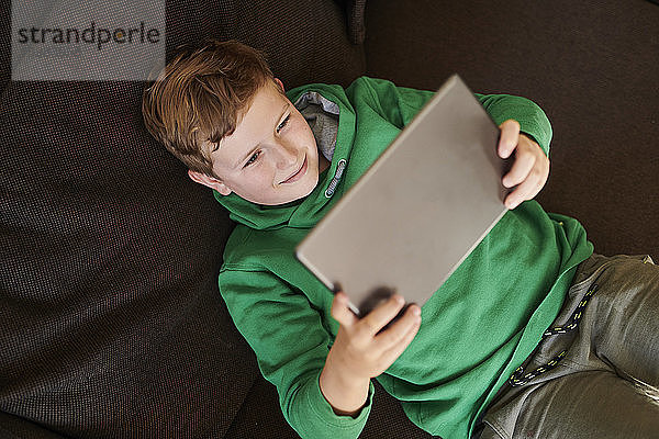 Lächelnder Junge  der ein digitales Tablet benutzt  während er zu Hause auf dem Sofa liegt