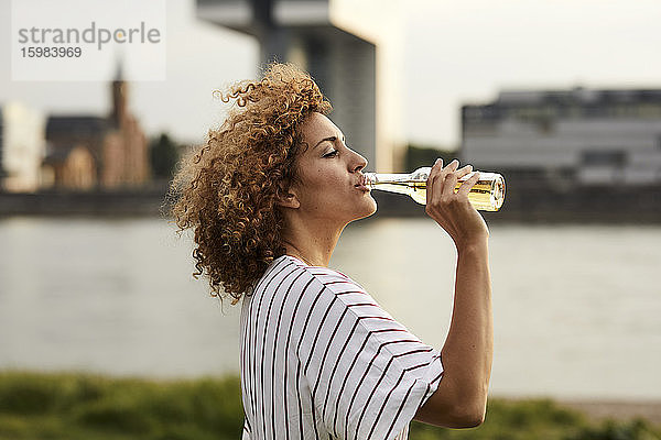 Porträt einer lächelnden Frau  die aus einer Glasflasche am Flussufer trinkt