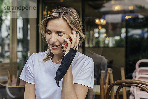 Porträt einer lächelnden Frau am Telefon mit einer Schutzmaske in der Hand vor einem geschlossenen Kaffeehaus