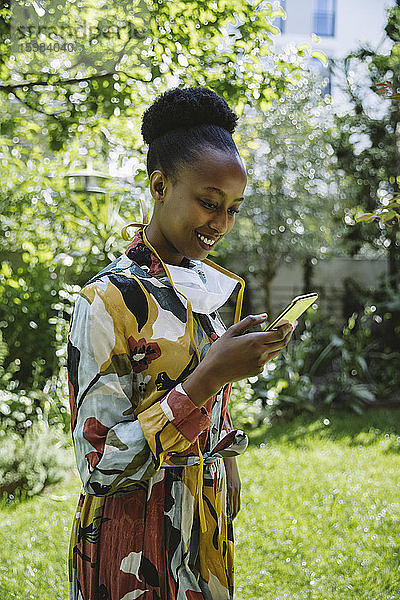 Porträt einer modischen jungen Frau mit Schutzmaske  die im Garten steht und auf ihr Mobiltelefon schaut