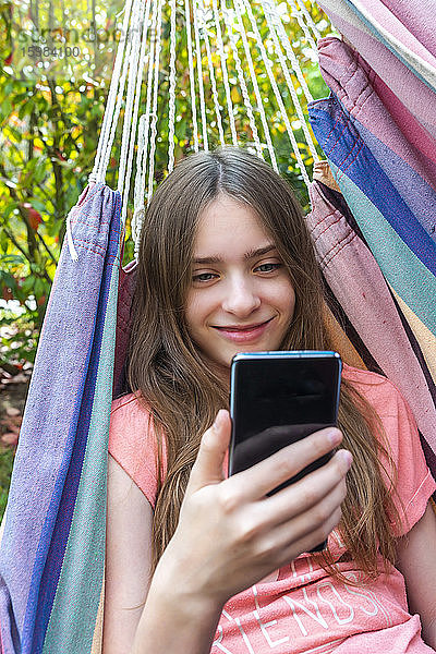 Porträt eines lächelnden Mädchens in der Hängematte  das auf sein Handy schaut