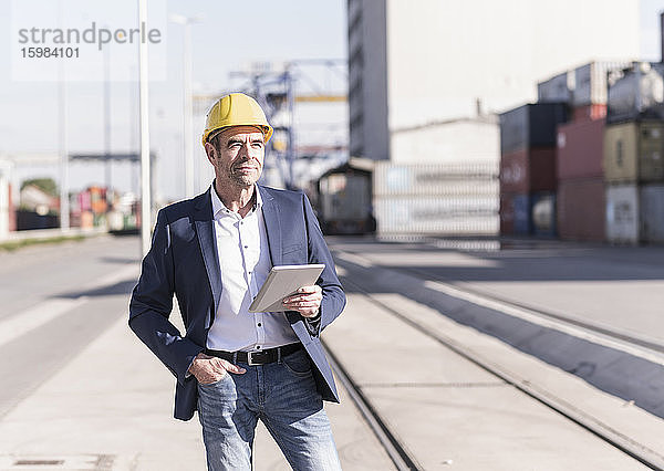 Porträt eines zufriedenen Geschäftsmannes mit Schutzhelm und digitalem Tablet auf einem Industriegelände