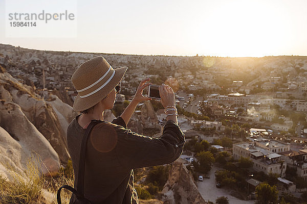 Seitenansicht einer jungen Frau  die während des Sonnenuntergangs mit ihrem Smartphone die Stadt fotografiert  Kappadokien  Türkei