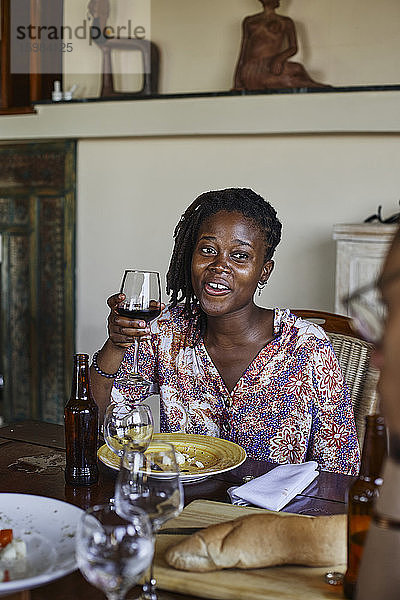 Lächelnde junge Frau mit Weinglas in der Hand  die während des Mittagessens am Esstisch sitzt