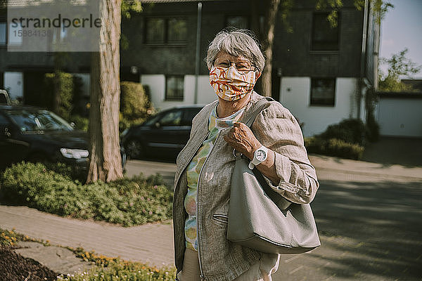 Ältere Frau trägt eine Gesichtsmaske  während sie an einem sonnigen Tag während des Ausbruchs der Pandemie im Freien steht