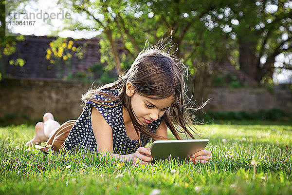 Lächelndes Mädchen  das auf einer Wiese im Garten liegt und ein digitales Tablet benutzt