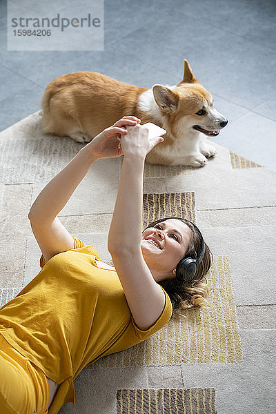 Frau mit Hund und Kopfhörern  die zu Hause auf dem Boden liegt und ihr Smartphone benutzt