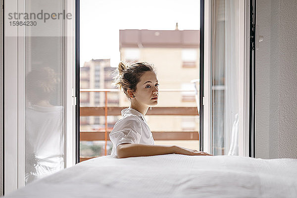 Seitenansicht einer nachdenklichen jungen Frau  die wegschaut  während sie zu Hause am Bett sitzt