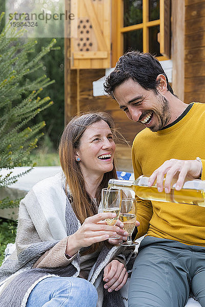 Glücklicher Mann gießt Wein in ein von seiner Freundin gehaltenes Glas  während er im Freien sitzt