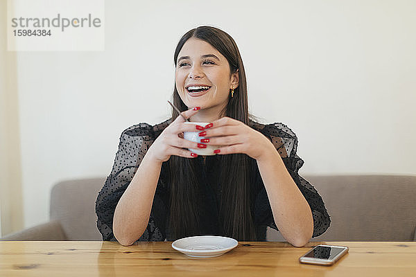 Fröhliche junge Frau hält eine Kaffeetasse  während sie am Tisch sitzt und in der Cafeteria wegschaut