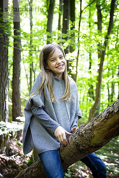 Porträt eines lächelnden niedlichen Mädchens  das auf einem Baumstamm im Wald sitzt