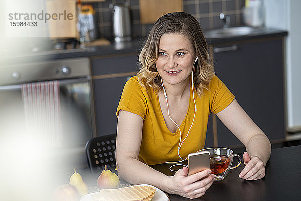 Frau mit Smartphone und Kopfhörern am Küchentisch zu Hause