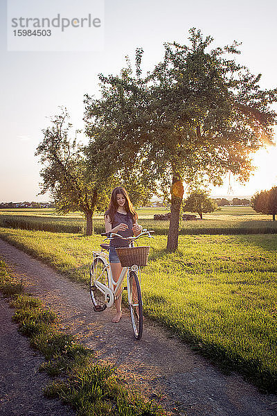 Teenager-Mädchen steht neben Fahrrad auf unbefestigten Weg bei Sonnenuntergang