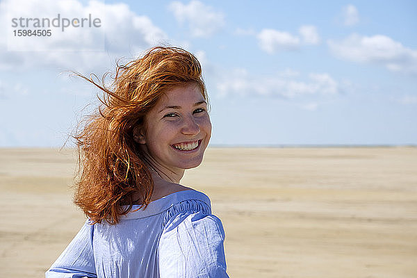 Lächelndes Teenager-Mädchen  das über die Schulter schaut  während es am Strand gegen den Himmel an einem sonnigen Tag steht