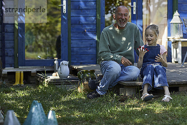 Glücklicher Großvater und Enkelin mit Himbeeren an den Fingerspitzen auf der Terrasse im Garten sitzend