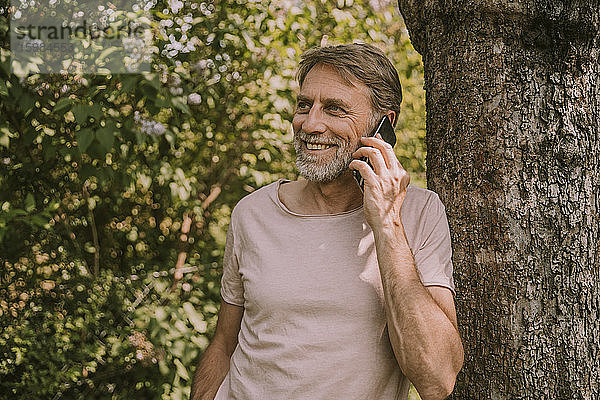 Lächelnder reifer Mann  der am Handy spricht  während er sich an einen Baum im Garten lehnt