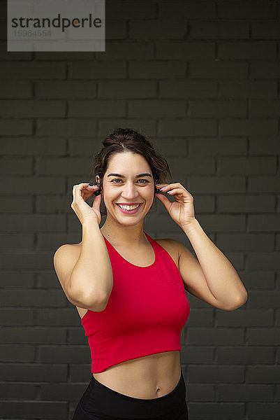 Lächelnde Sportlerin  die Musik über drahtlose Kopfhörer hört  während sie an einer Mauer steht