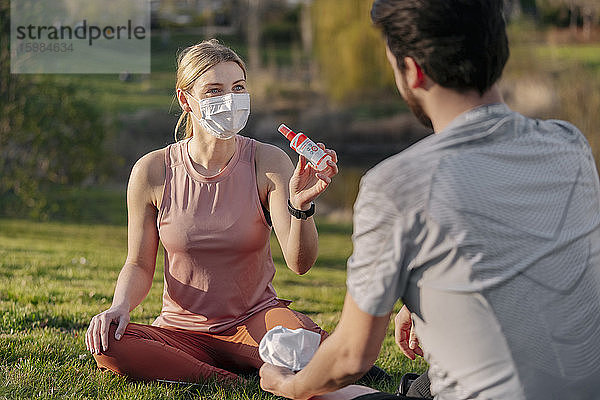 Frau mit Gesichtsmaske zeigt Mann Handdesinfektionsmittel  während er im Park sitzt