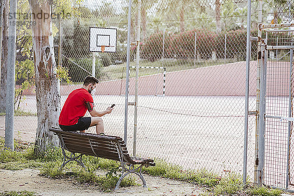 Mittelgroßer erwachsener Mann mit Gesichtsmaske  der sein Smartphone benutzt  während er auf einer Bank im Park sitzt