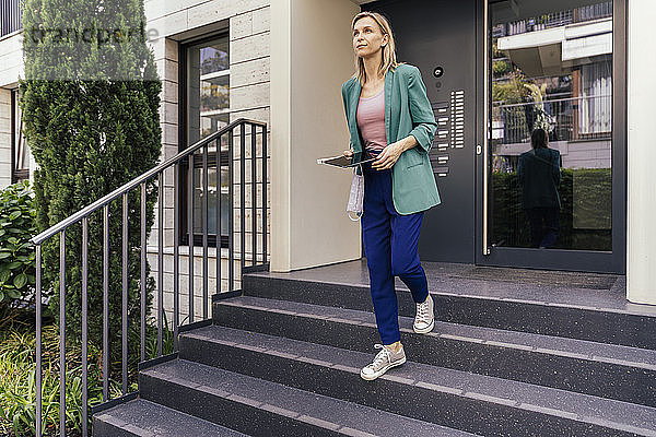 Immobilienmakler mit digitalem Tablet und Gesichtsmaske in der Hand geht die Treppe eines Hauses hinunter