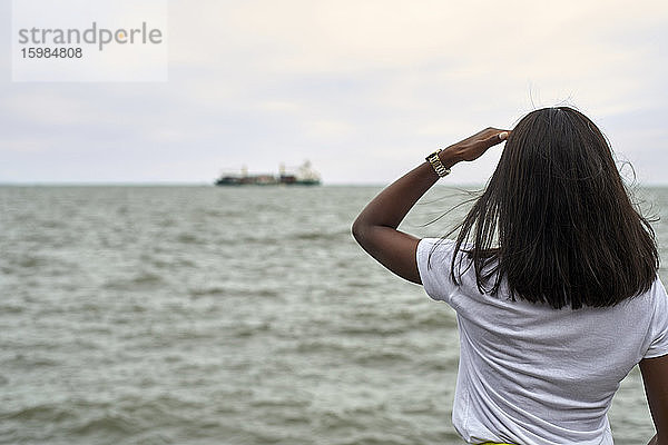 Rückenansicht einer jungen Frau vor dem Meer mit Blick auf ein Schiff am Horizont