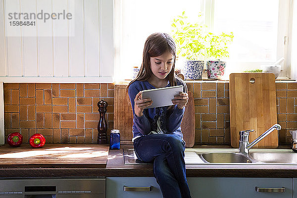 Mädchen sitzt am Waschbecken in der Küche und schaut auf ein digitales Tablet