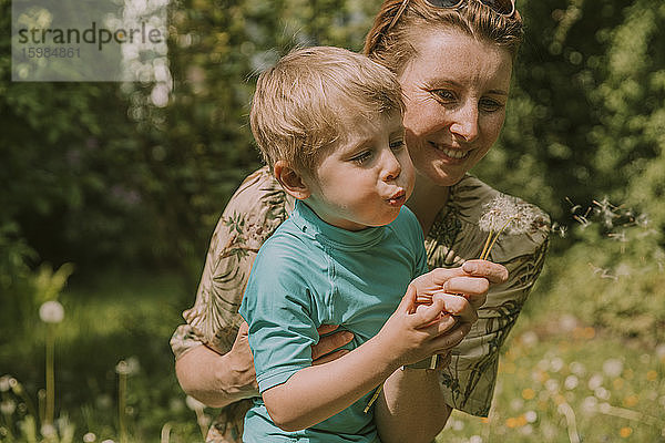 Glückliche Frau mit Sohn  der an einem sonnigen Tag Löwenzahnsamen im Garten ausbläst