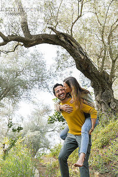 Fröhlicher Mann  der seine romantische Freundin huckepack nimmt  während er auf dem Lande gegen Bäume steht