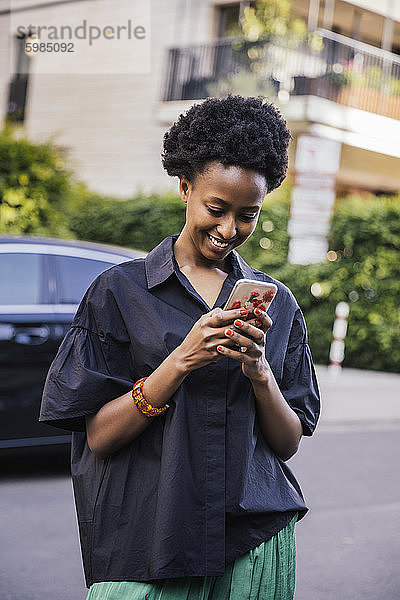 Porträt einer lächelnden jungen Frau  die auf der Straße steht und auf ihr Mobiltelefon schaut