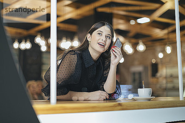 Lächelnde schöne junge Frau  die ihr Smartphone hält und in ein beleuchtetes Café schaut