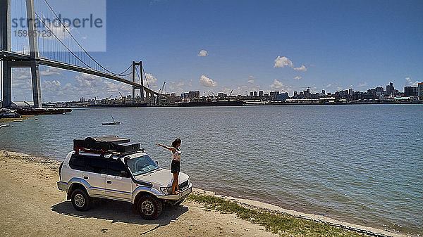 Mosambik  Katembe  Erwachsene Frau steht mit erhobenen Armen auf der Stoßstange eines Geländewagens und bewundert die Aussicht auf die Maputo-Bucht mit der Stadt und der Maputo-Katembe-Brücke im Hintergrund