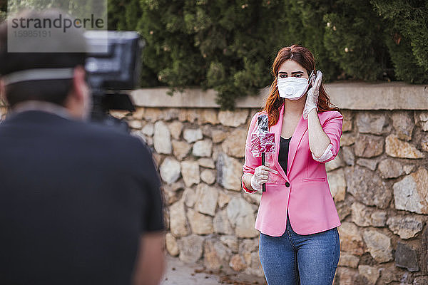 Kameramann filmt Reporter mit Mikrofon und Maske  während er an der Wand steht