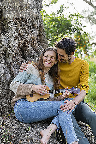 Liebender Mann umarmt glückliche Freundin  die Gitarre spielt  während er gegen einen Baumstamm sitzt