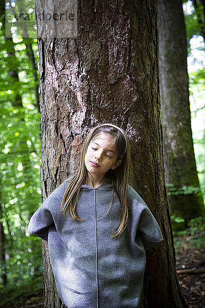 Junges Mädchen mit geschlossenen Augen an einem Baumstamm im Wald stehend