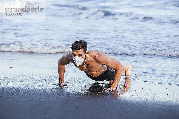 Shirtless Mitte erwachsenen Mann trägt Gesichtsmaske  während tun Push-ups am Strand bei Sonnenuntergang