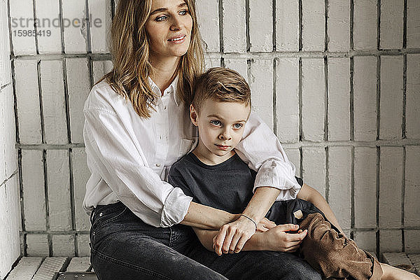 Nachdenkliche blonde Frau  die sich umarmt  während sie mit ihrem Sohn auf einer Nische zu Hause sitzt