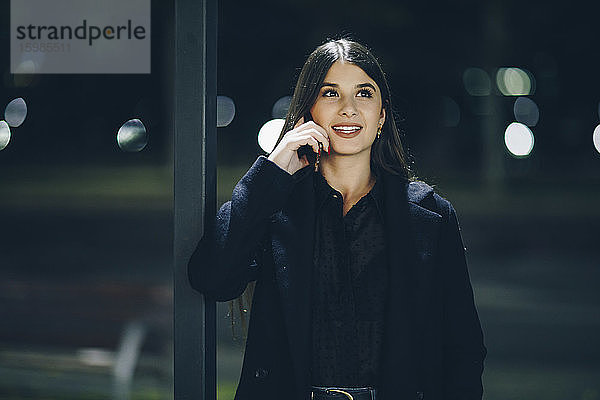 Lächelnde schöne junge Frau  die wegschaut  während sie nachts im Freien mit dem Handy telefoniert