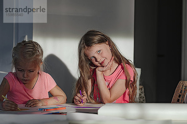 Glückliches Mädchen  das mit seiner Schwester sitzt und zu Hause auf Papier malt