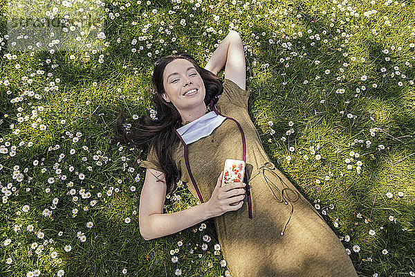 Frau mit heruntergelassener Gesichtsmaske genießt ihre Freizeit  während sie im Gras mit Gänseblümchen liegt