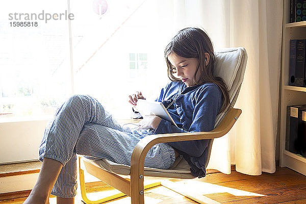Junges Mädchen sieht sich ein Online-Video auf einem digitalen Tablet an  während sie zu Hause am Fenster sitzt