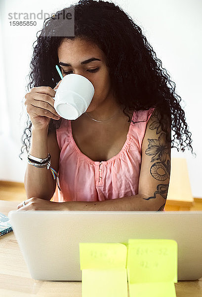 Junge Frau arbeitet von zu Hause aus mit Laptop und trinkt Kaffee