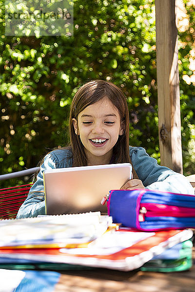 Mädchen sitzt am Gartentisch  macht Hausaufgaben und benutzt ein Tablet